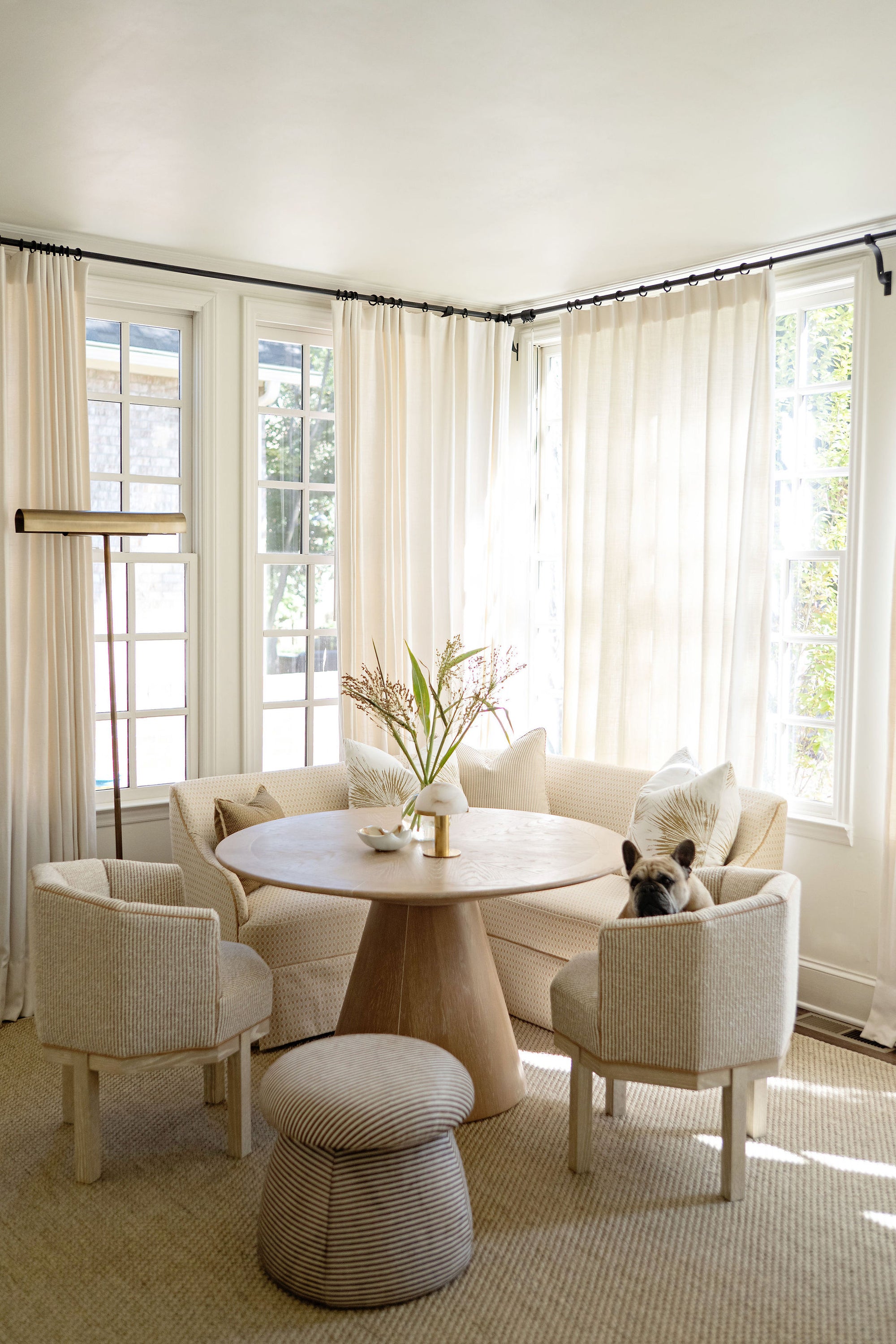 A bright, sunlit corner dining room featuring a round wooden pedestal table, two light beige upholstered armchairs, a matching round pouf, and a curved sofa. A small dog is sitting inside one of the armchairs, and the room is decorated with sheer white curtains and natural light.
