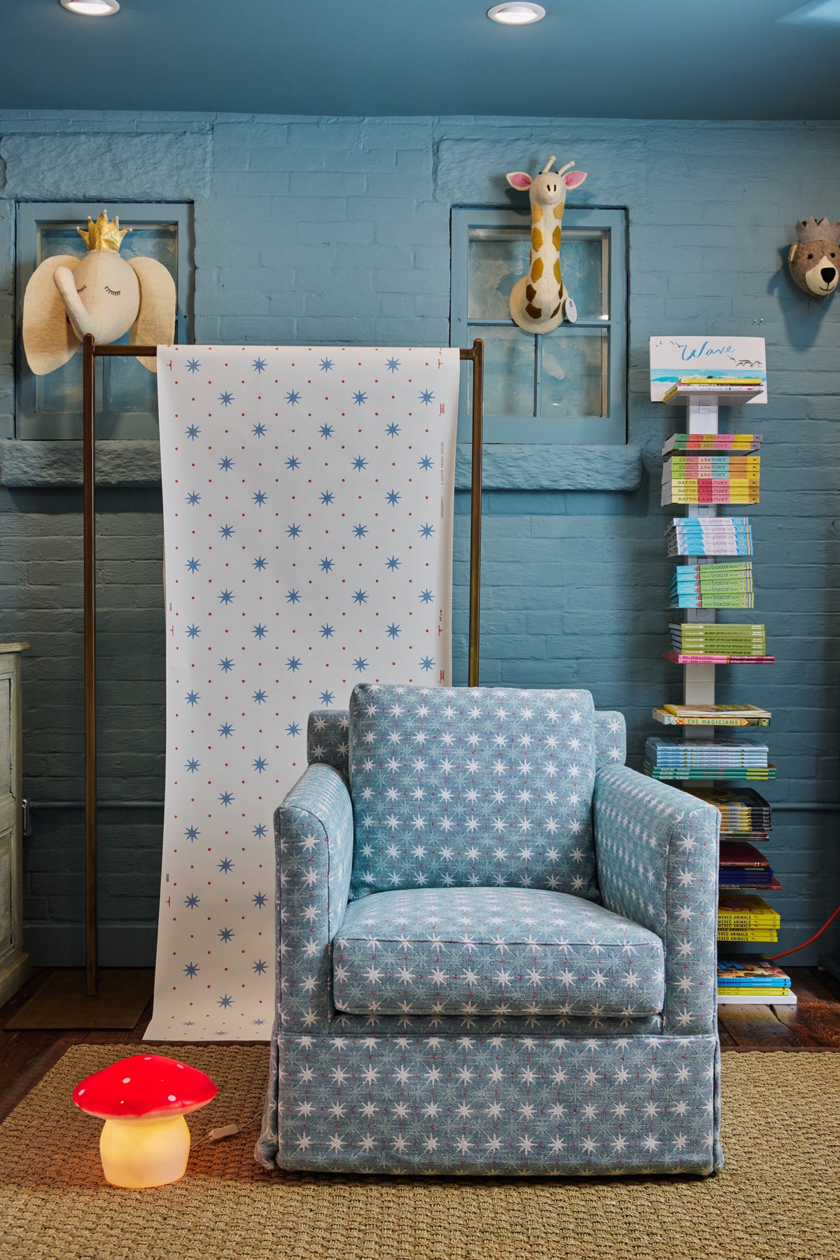 A blue and white patterned upholstered armchair in a styled room setting with a matching patterned fabric backdrop hanging on a stand, blue brick wall, bookshelf, and decorative wall pieces.