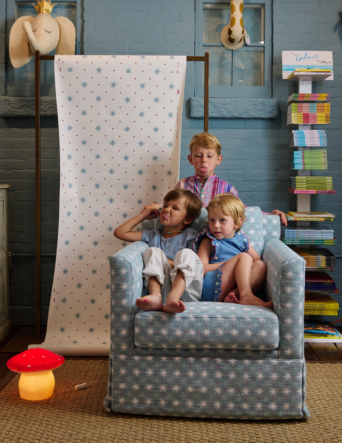 A blue upholstered armchair with a white star pattern is shown in a styled room with three children sitting on it. A matching star-patterned fabric panel hangs on a stand behind the chair, and the room includes blue walls, books, and decorative objects.
